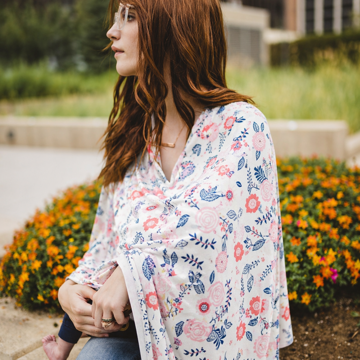 Woman in a floral baby nursing cover sitting on a stone bench with flowers and buildings in the background