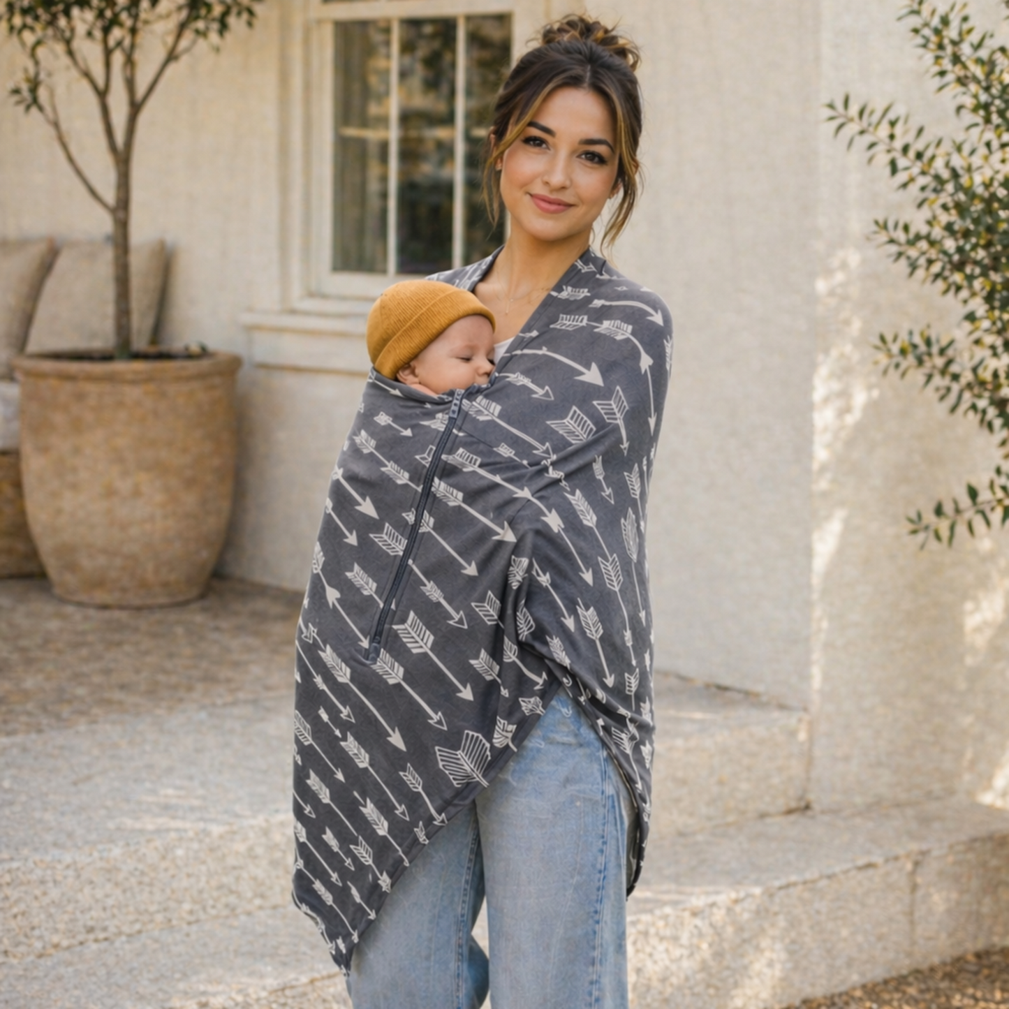 Woman holding a baby wrapped in a gray patterned carrier cover outdoors.