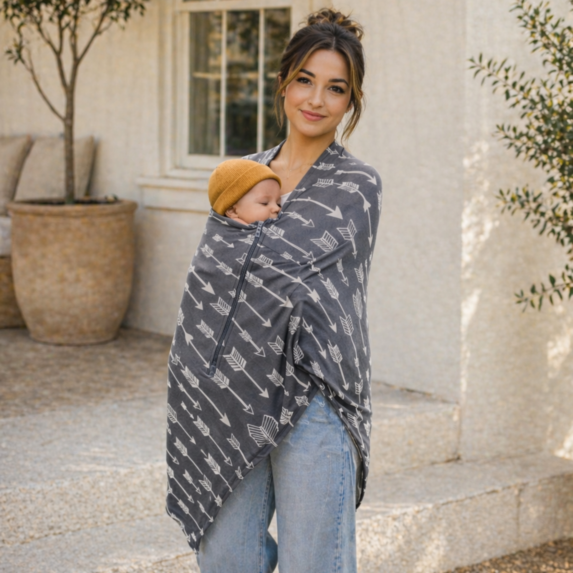 Woman holding a baby wrapped in a gray patterned carrier cover outdoors.