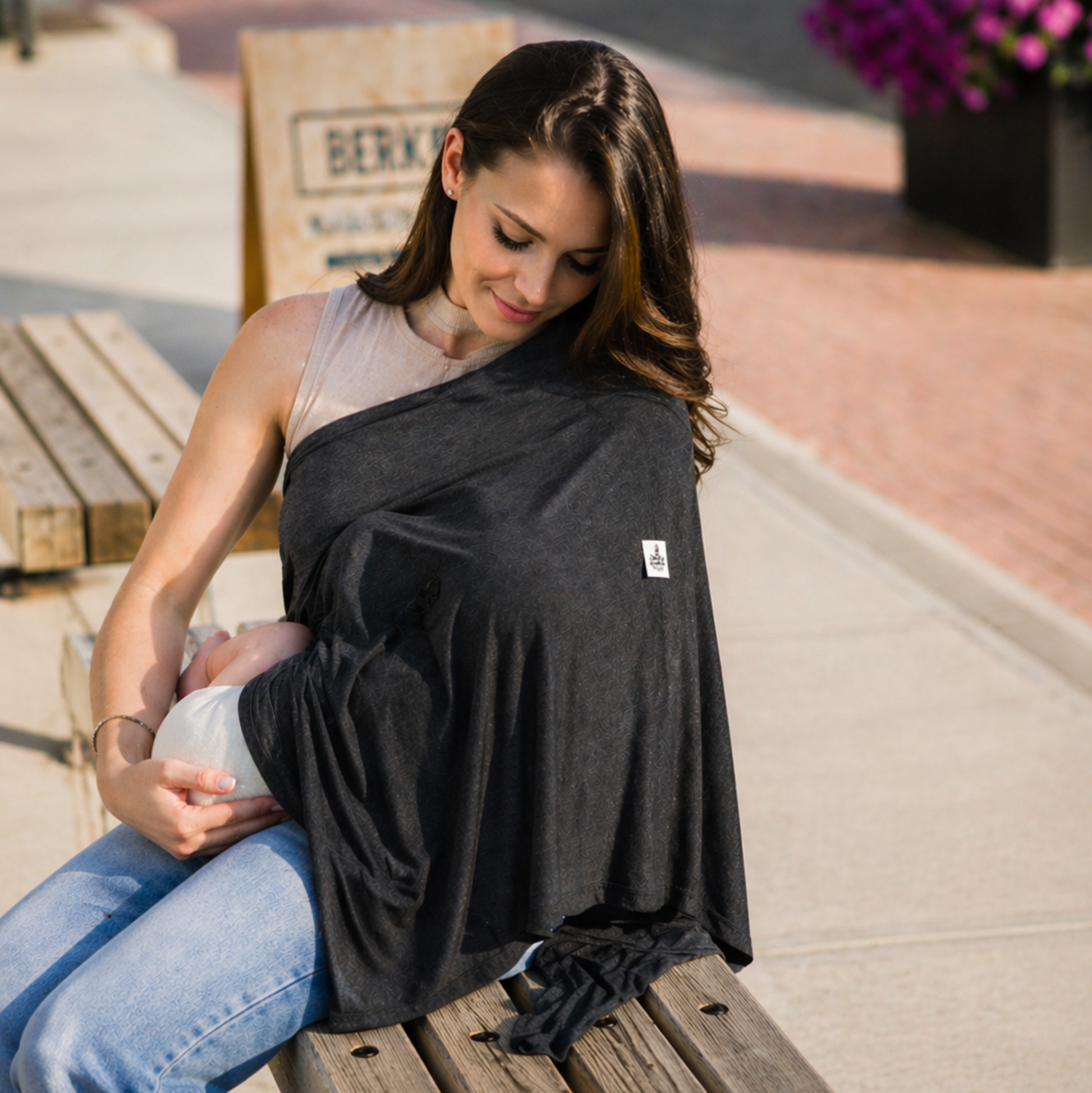 Woman sitting on a bench wearing a dark gray baby carrier with a baby inside, on a sidewalk.