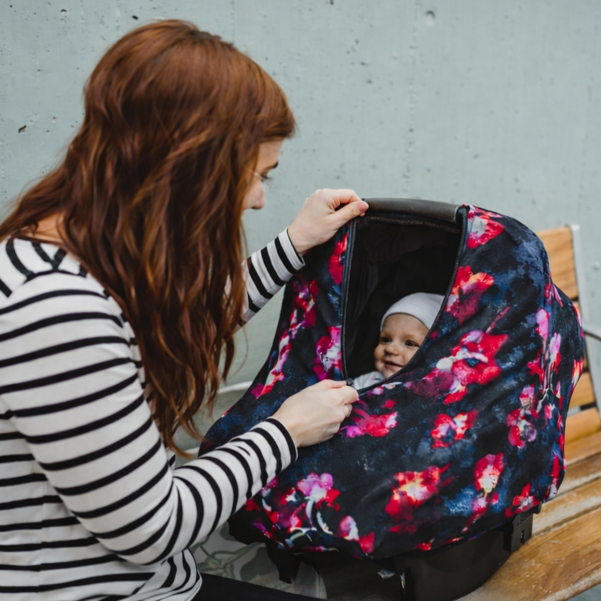 Woman with a baby in a floral car seat cover on a bench.